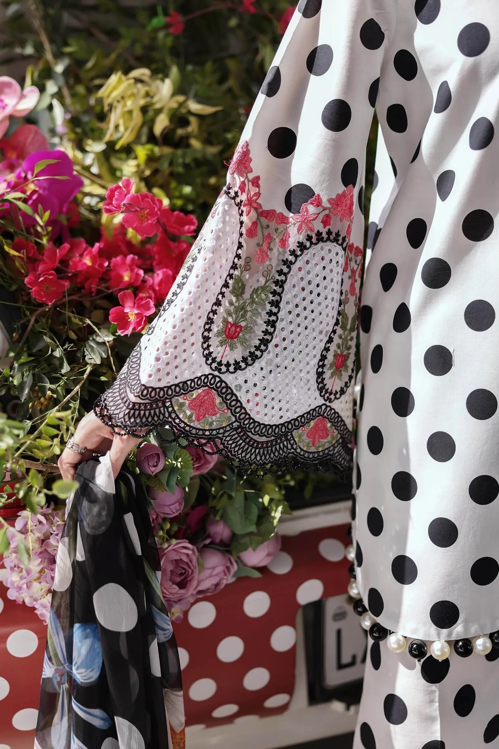 White polka dot dress with floral embroidery and lace trim, styled with pearls against vibrant garden blooms