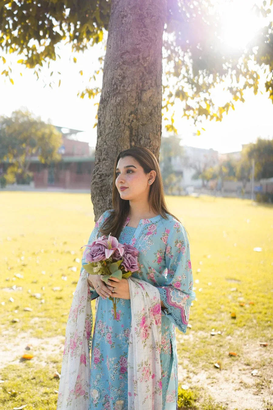 Woman in floral blue kurta with dupatta, holding roses outdoors under tree